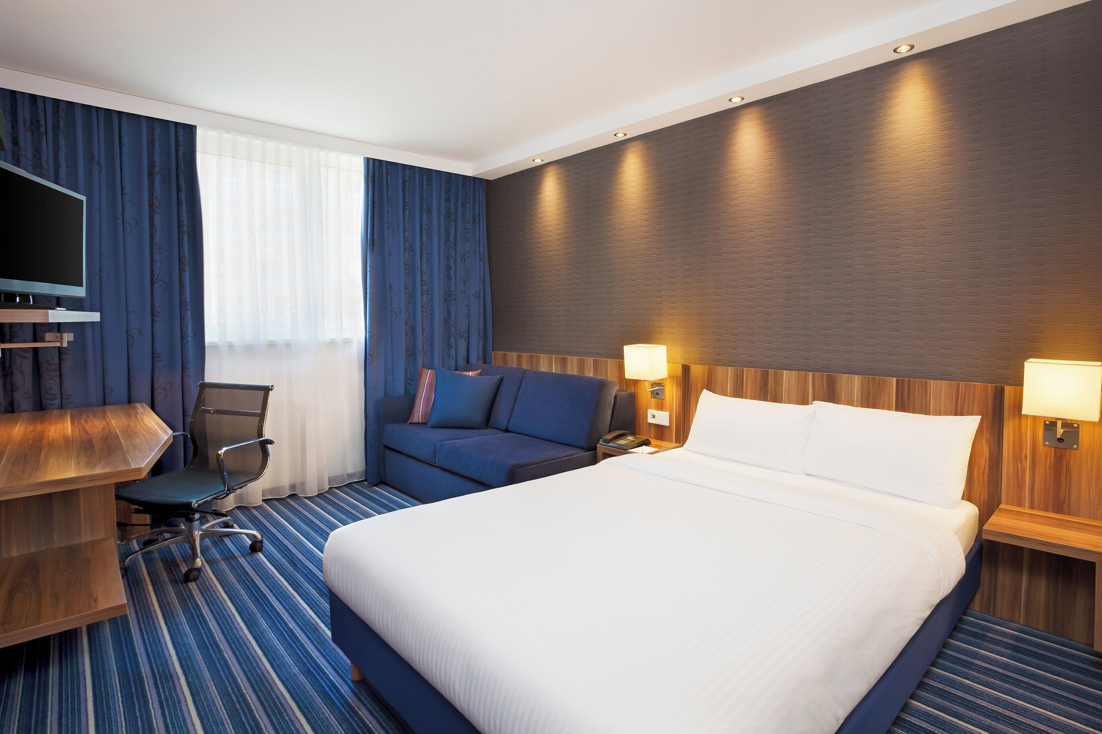 A hotel room with a large bed, a desk with a flat-screen TV, a blue sofa, and a wooden headboard.