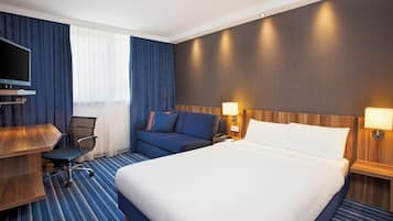 A hotel room with a large bed, a desk with a flat-screen TV, a blue sofa, and a wooden headboard.
