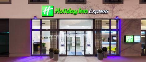 Exterior de un hotel Holiday Inn Express con fachada de vidrio, una zona de recepción y un letrero digital que muestra el nombre del hotel.