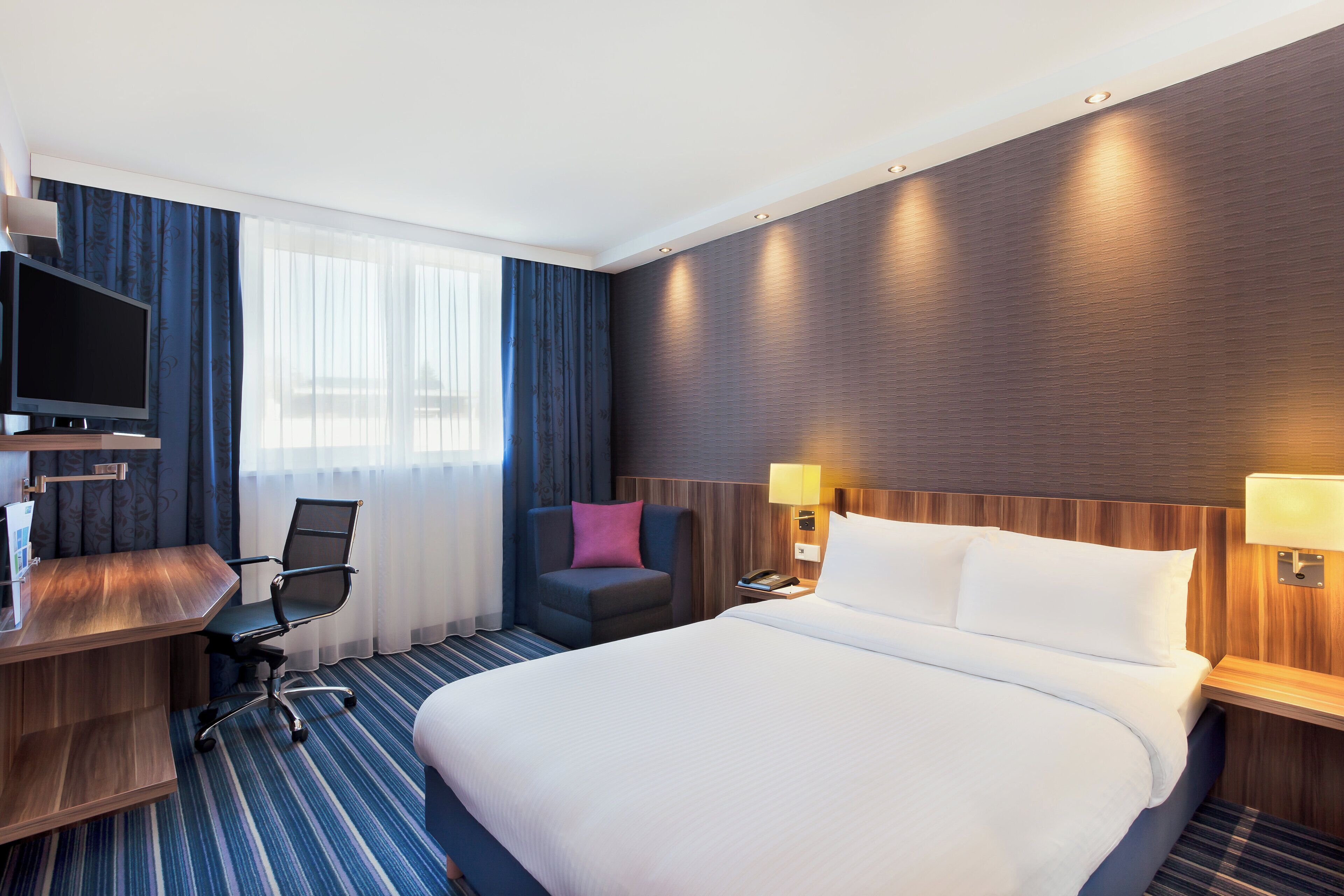 Une chambre d’hôtel avec un grand lit, un bureau avec un ordinateur, une chaise, une télévision et une table de chevet avec une lampe.