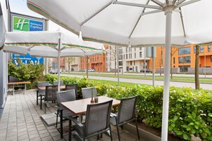 Un área de estar al aire libre con mesas y sillas bajo sombrillas, un letrero de Holiday Inn Express y vistas a una carretera y edificios al fondo.