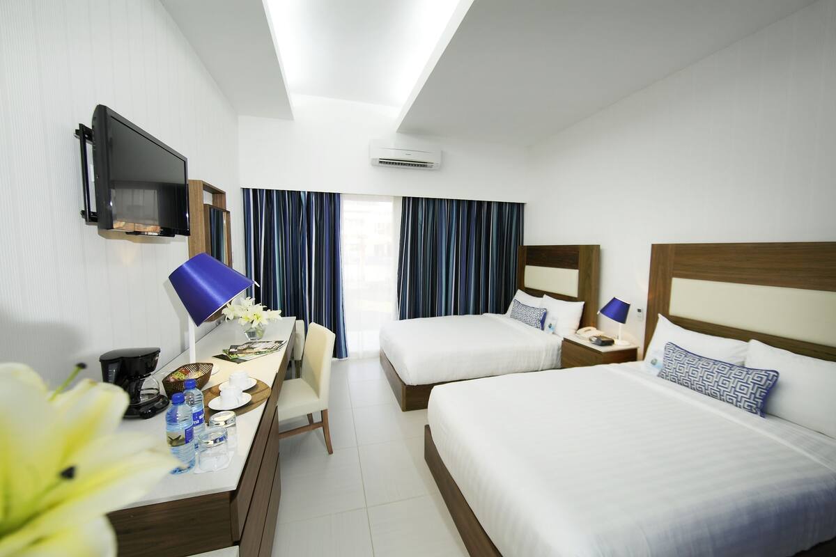 A hotel room with two beds, a desk with a chair, a TV mounted on the wall, and an air conditioning unit.