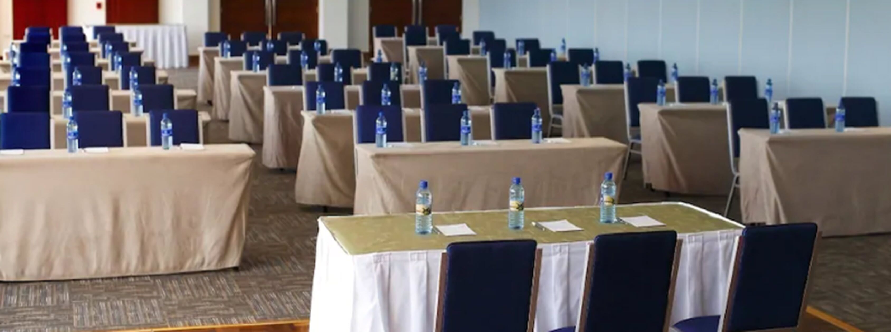 Uma sala de conferência com fileiras de mesas e cadeiras, cada mesa com uma garrafa de água e papéis.
