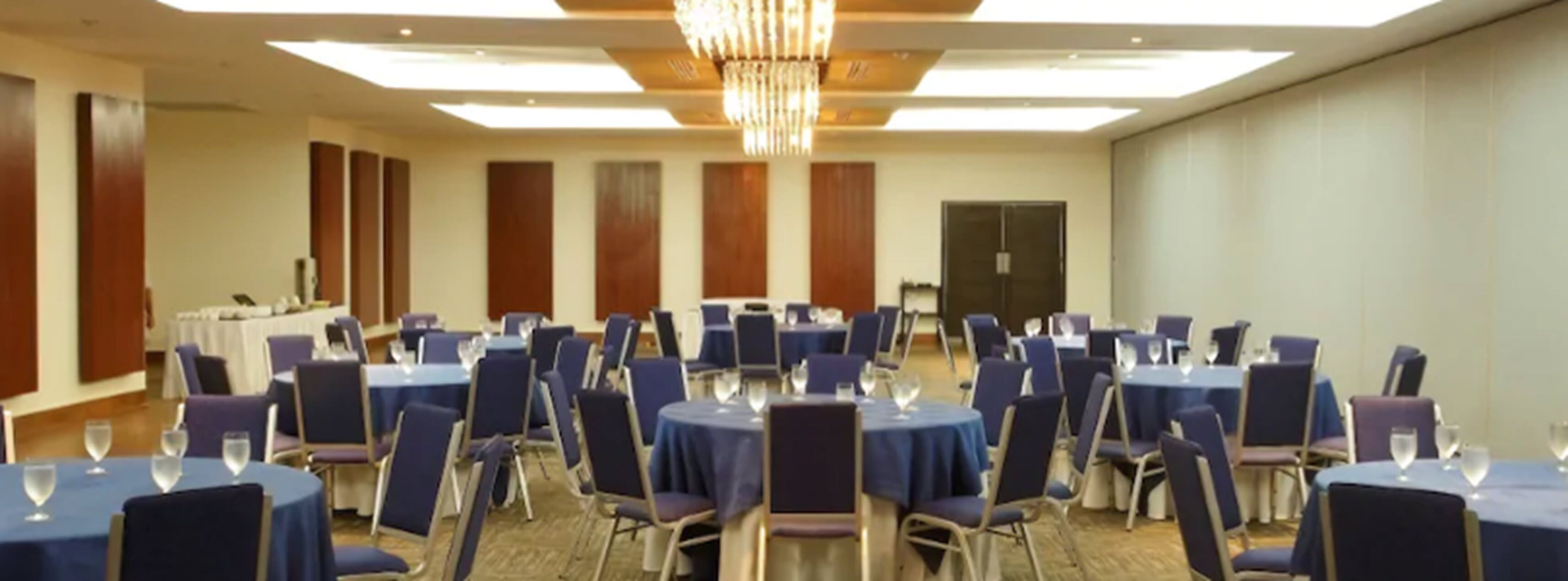 Een congrescentrum met ronde tafels en stoelen, een kroonluchter, en een grote deur op de achtergrond.