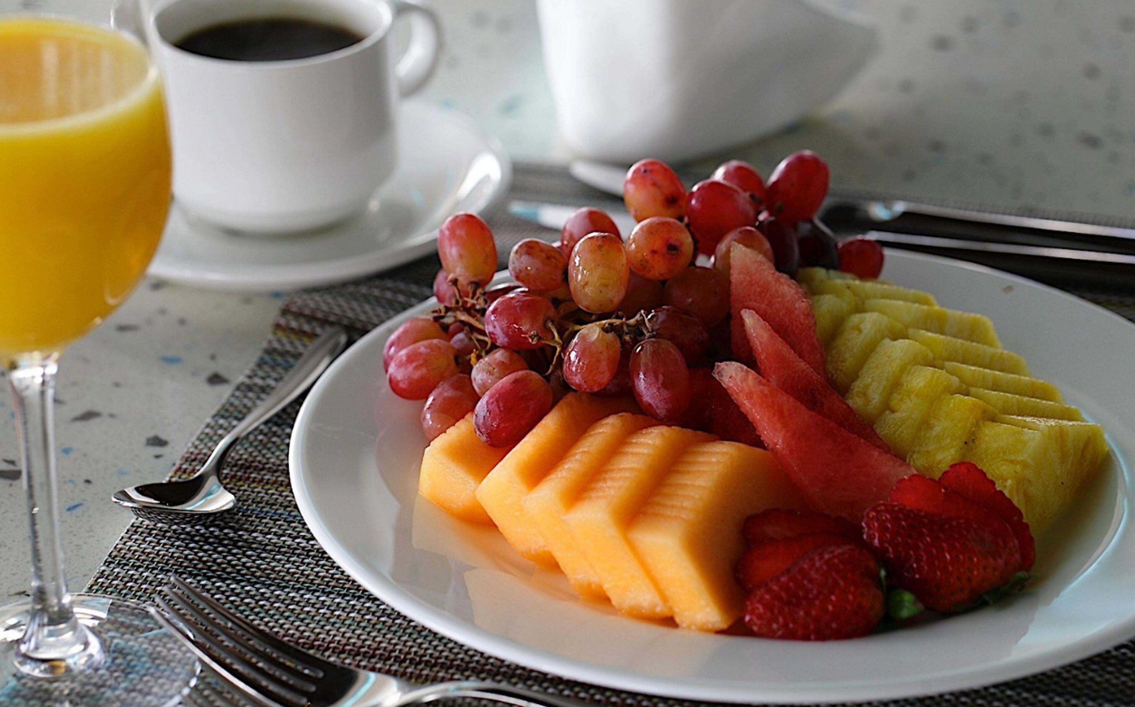 Un plato de fruta que incluye uvas, sandía, melón y fresas, con un vaso de jugo de naranja y una taza de café sobre una mesa.