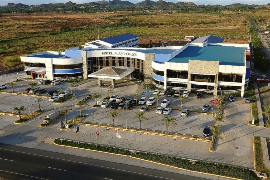 Un hotel con un edificio central y dos alas, un estacionamiento con varios autos, y un paisaje circundante.