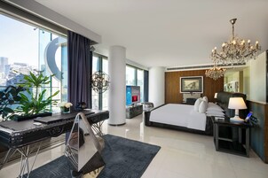 A modern hotel room with a large bed, a desk with a computer, a desk lamp, a chandelier, and a view of the city.