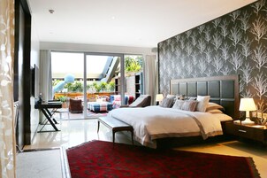 A modern bedroom with a large bed, a red rug, a nightstand, and a view of a balcony with plants.