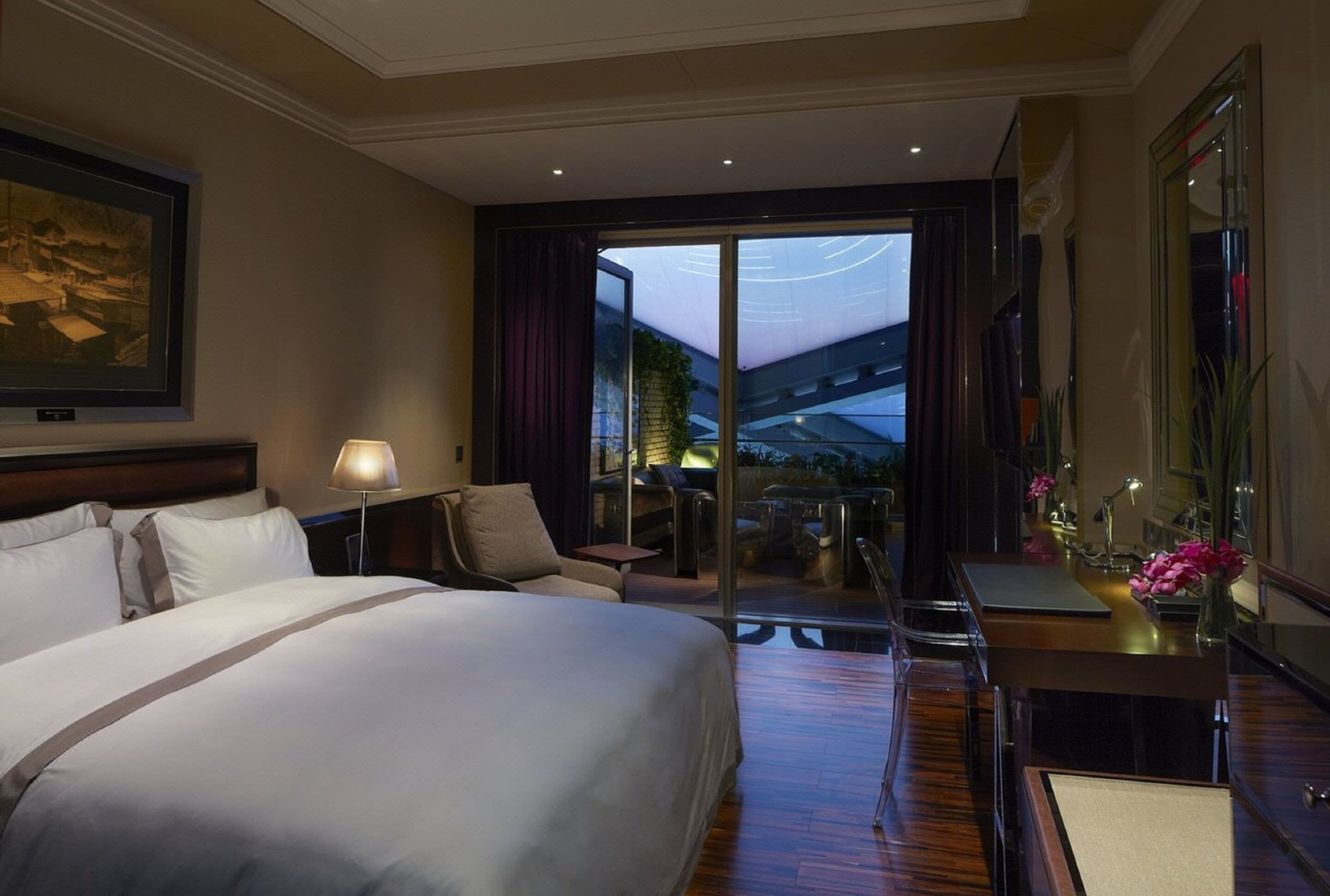 A hotel room with a large bed, a desk, a chair, a balcony with a view, and a painting on the wall.
