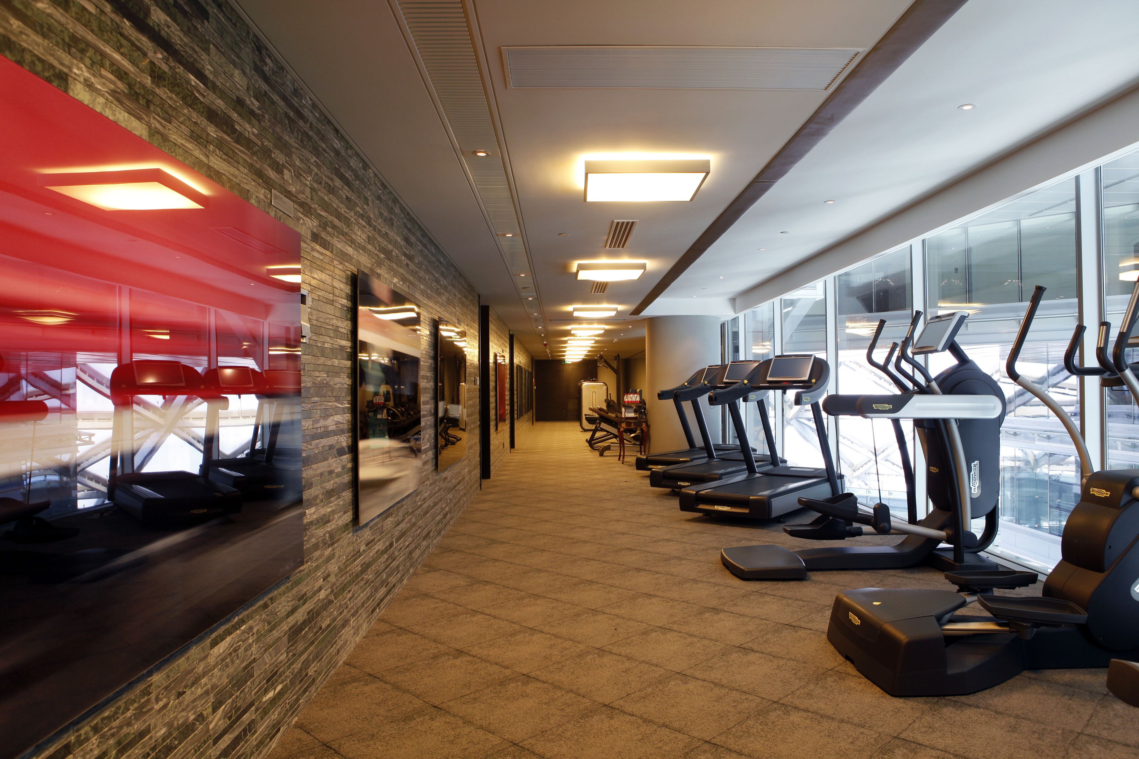 A gym with treadmills and exercise equipment, a brick wall, and large windows.