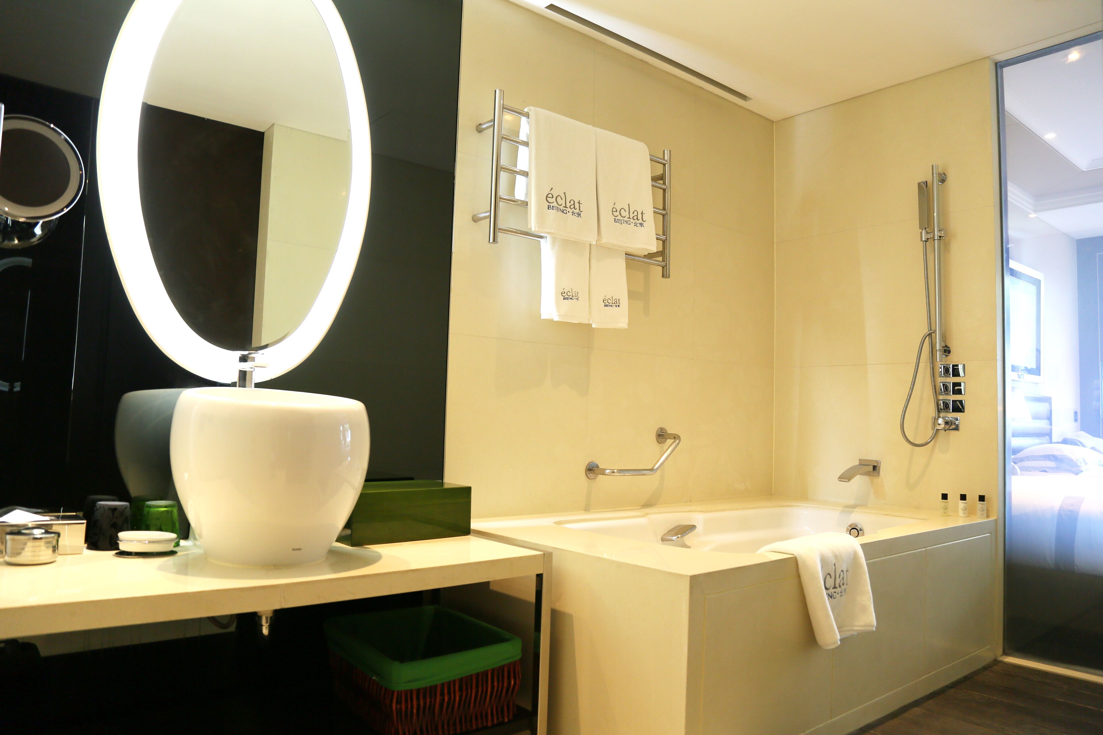 Un baño moderno con un espejo ovalado grande, un lavamanos blanco, una bañera y una ducha. Hay dos toallas colgadas en un perchero y un cubo de basura verde debajo del lavamanos.