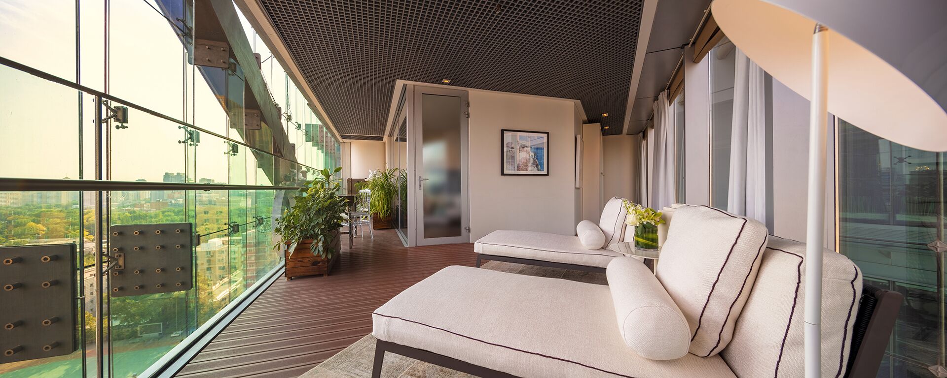 A modern balcony with a glass railing, a wooden floor, and a sofa with cushions.
