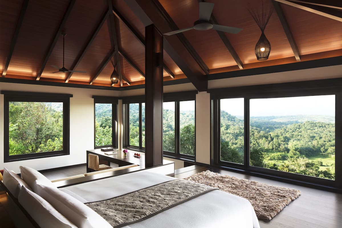 A bedroom with a large bed, a desk, and a view of a forest through large windows.