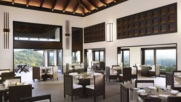 A spacious dining area with high ceilings and large windows offering mountain views. Tables are set with white tableware and chairs are upholstered in dark fabric.