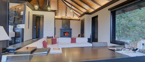 A modern kitchen with a large island, a seating area with a fireplace, and a view through a window.