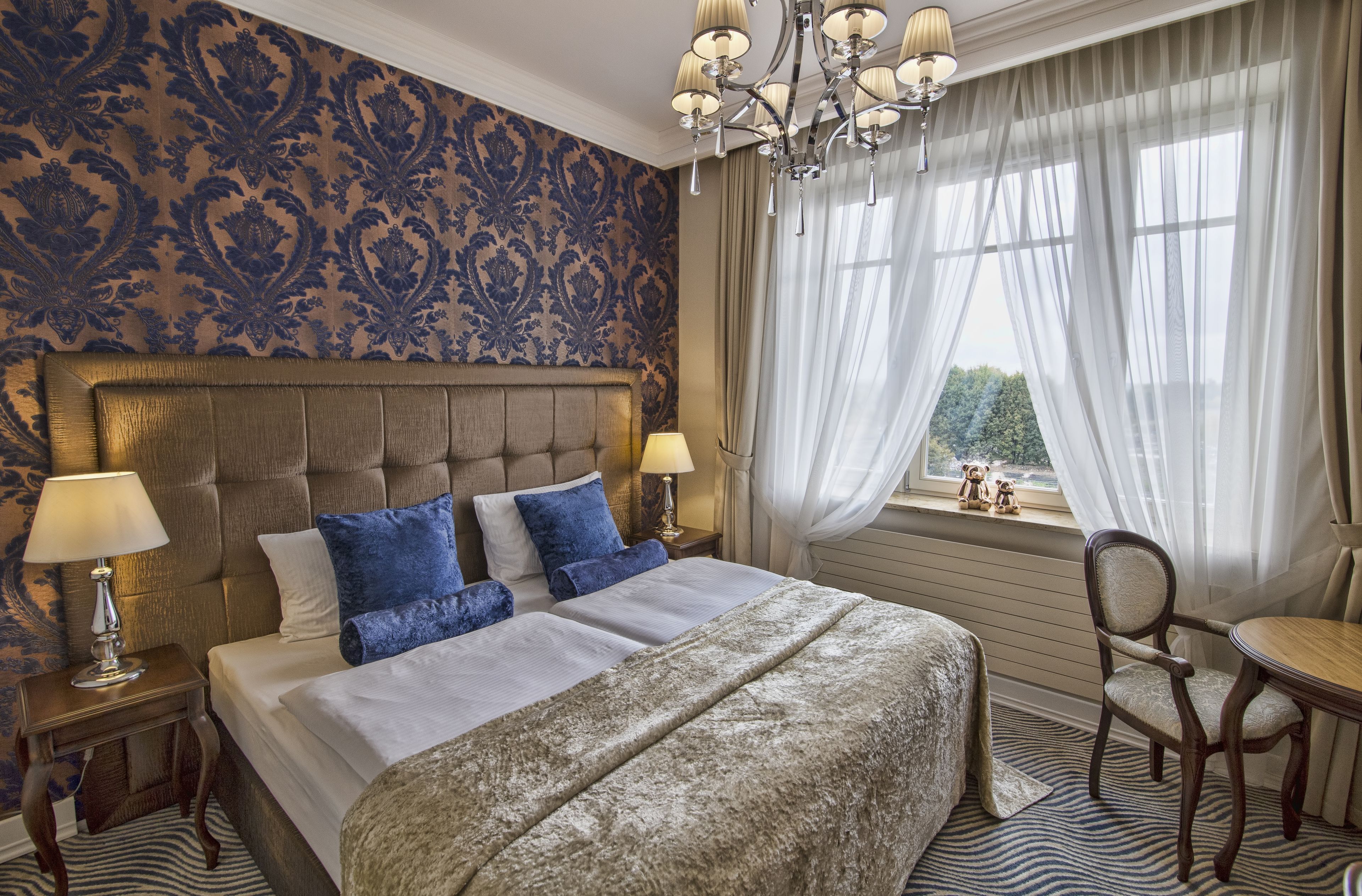 A bedroom with a large bed, two bedside tables with lamps, a chandelier, a window with sheer curtains, and a view of greenery outside.