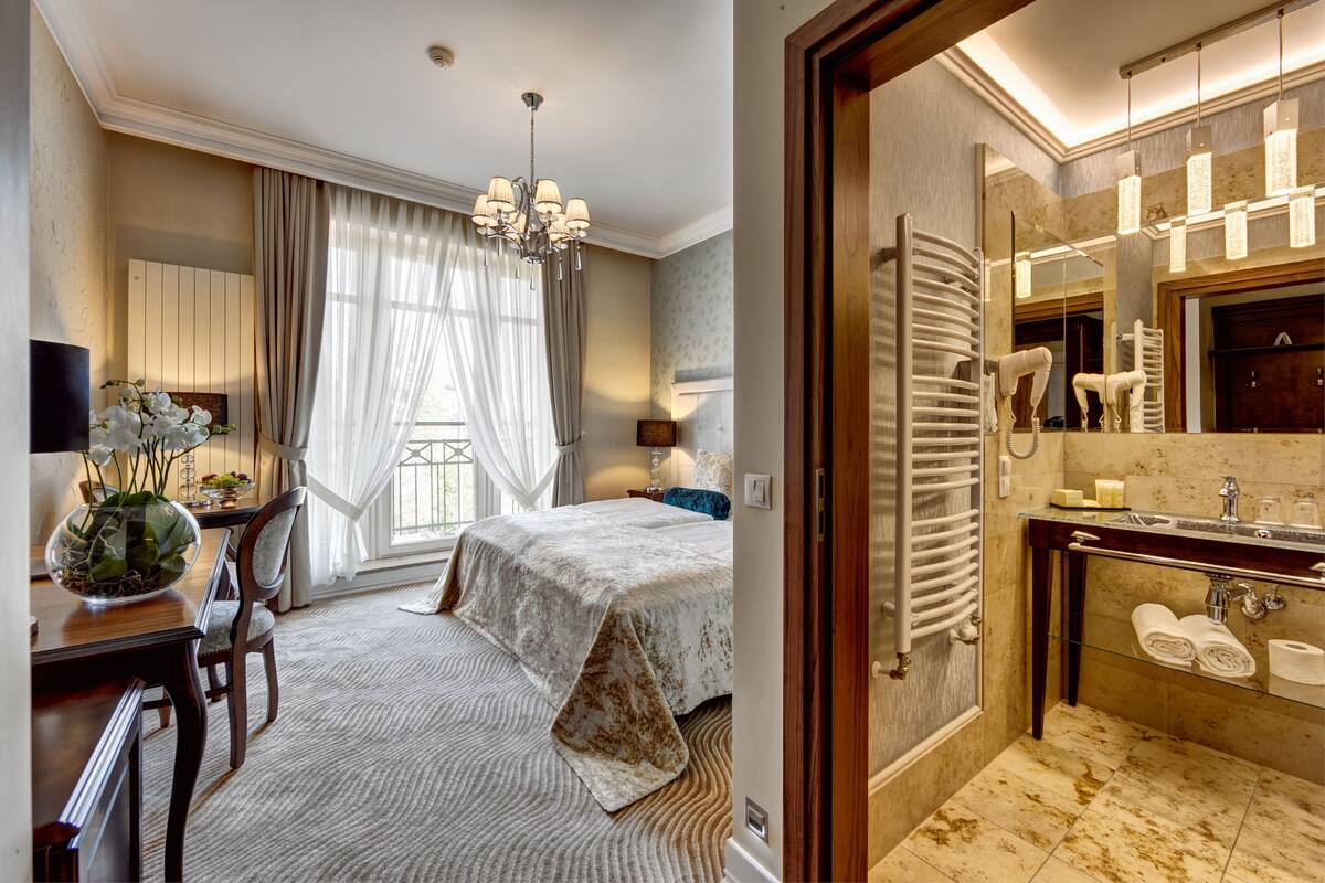 Hotelkamer met een bed, bureau, stoel, een kroonluchter, groot raam met gordijnen en een badkamer met spiegel, wastafel en handdoekhouder.