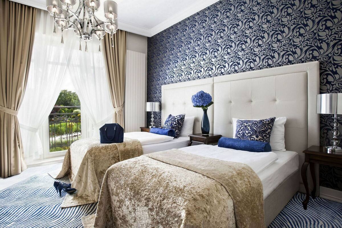 Een hotelkamer met twee bedden, een kroonluchter, uitzicht op groen en een behang met patroon.