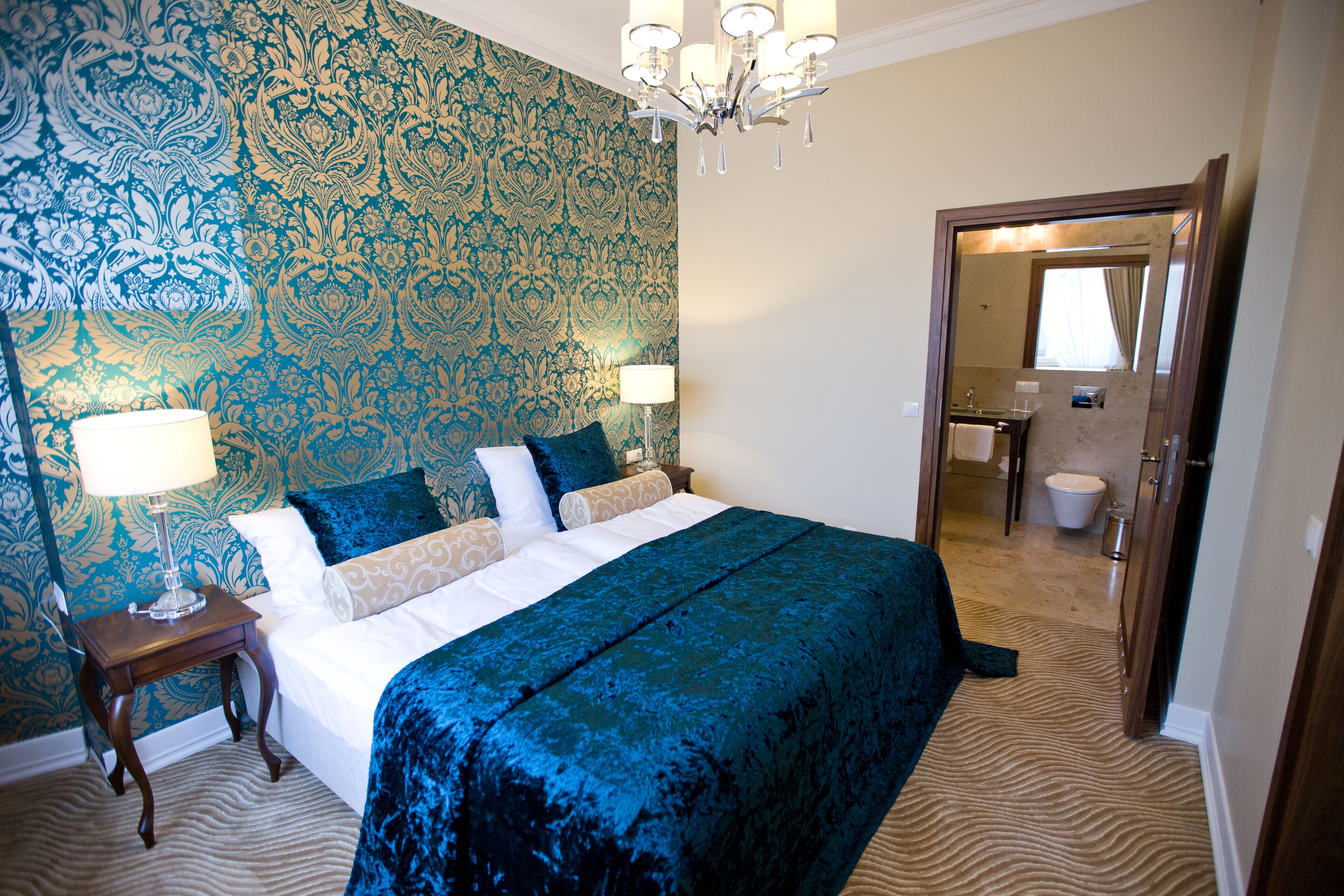 Une chambre d’hôtel avec un grand lit, deux tables de chevet, un lustre et un miroir qui reflète la salle de bain.