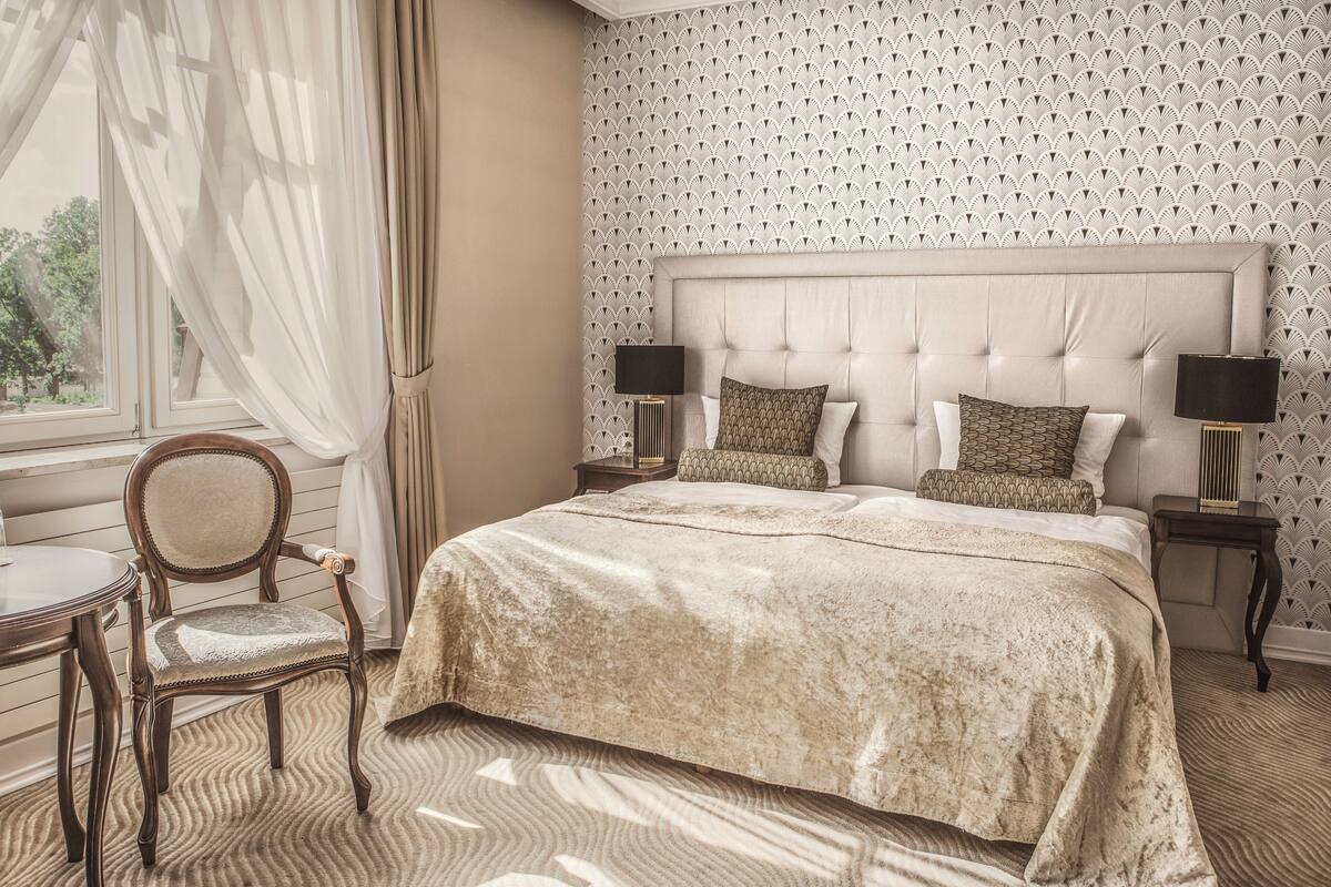 Een slaapkamer met een groot bed, twee nachtkastjes, een stoel en een raam met gordijnen.