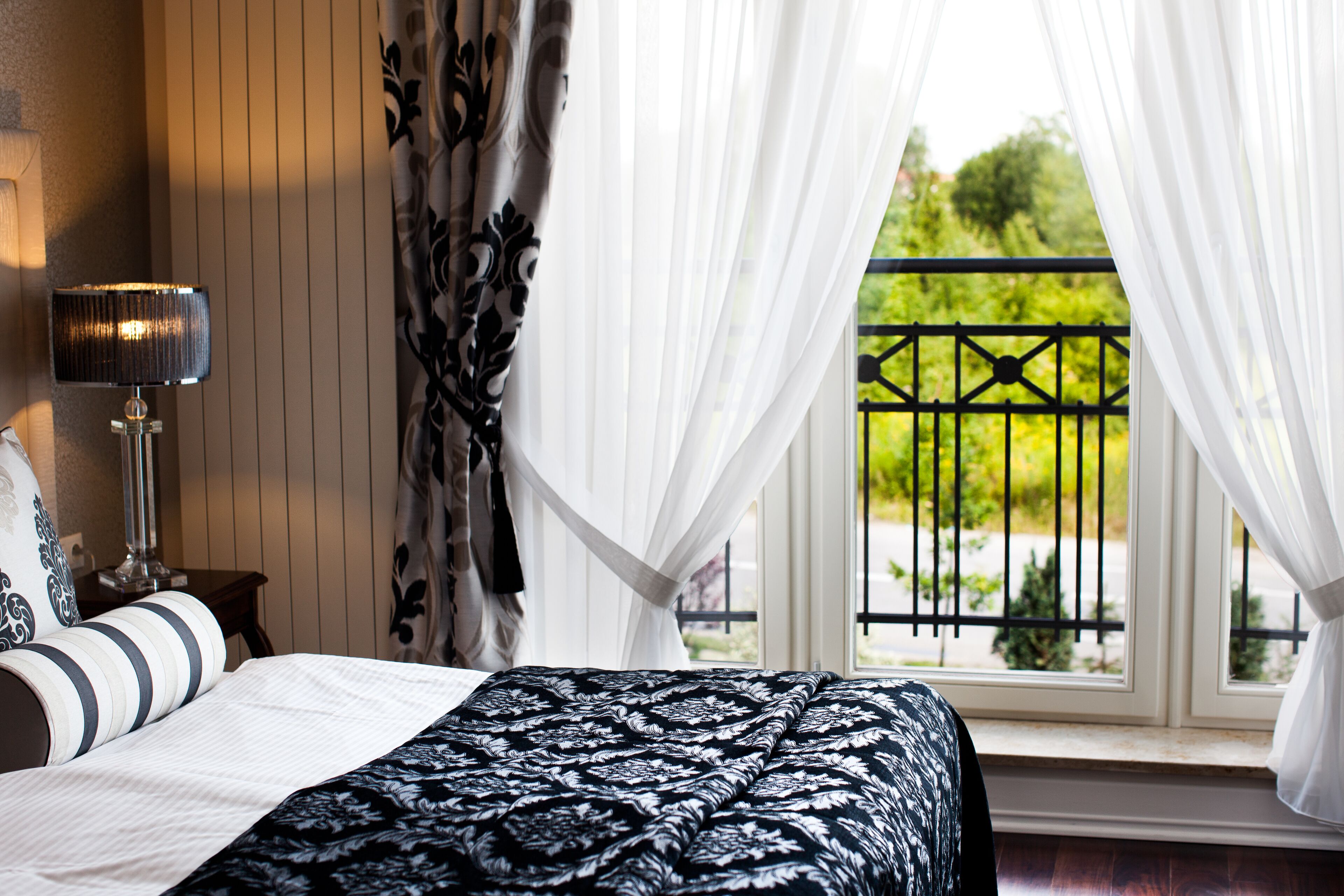 Ein Schlafzimmer mit einem Bett, einer Lampe, einem Fenster mit Vorhängen und Blick ins Grüne.