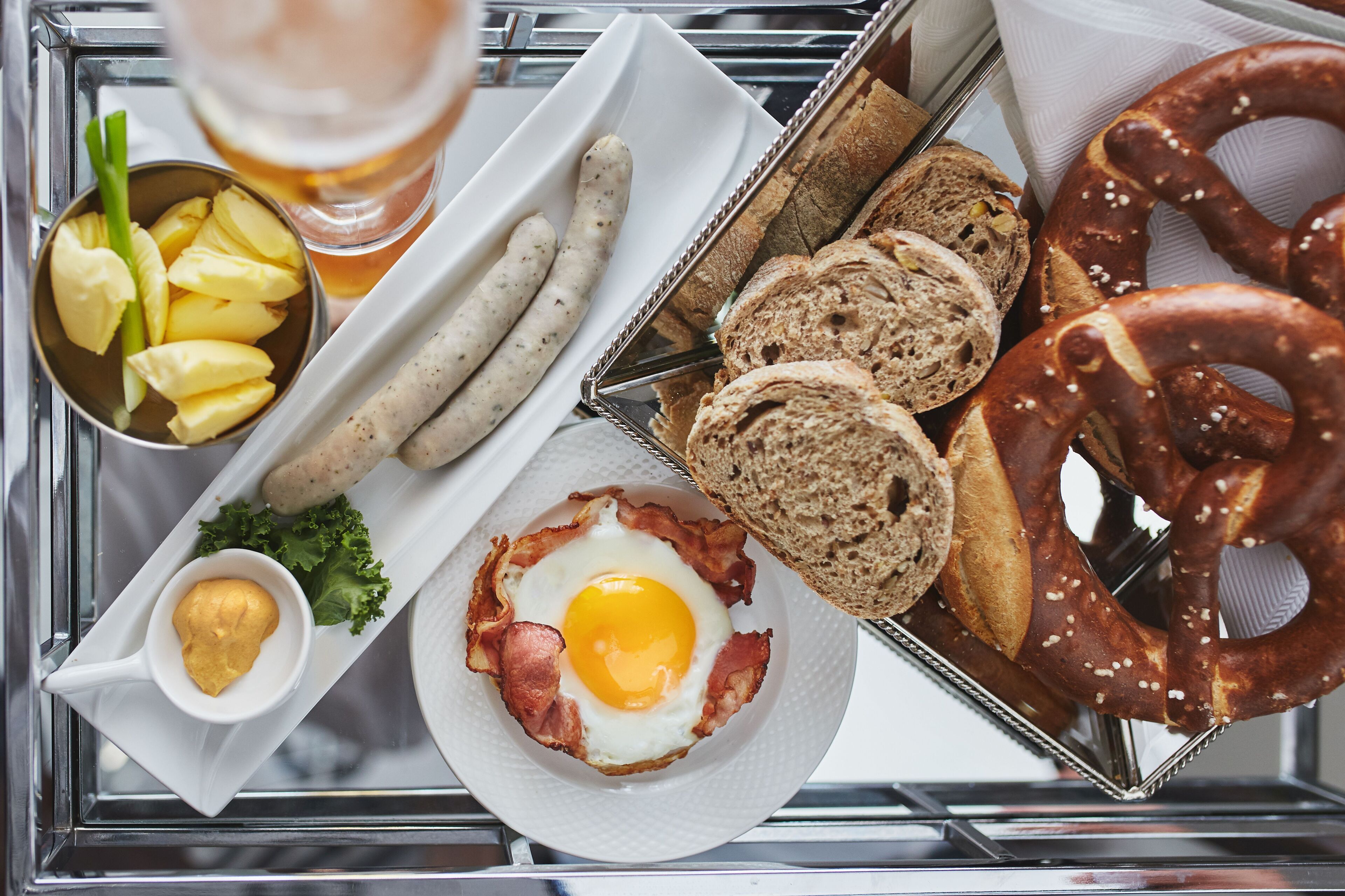 Una bandeja con alimentos para el desayuno, como salchichas, pretzels, pan, un huevo frito y un recipiente pequeño de mostaza.