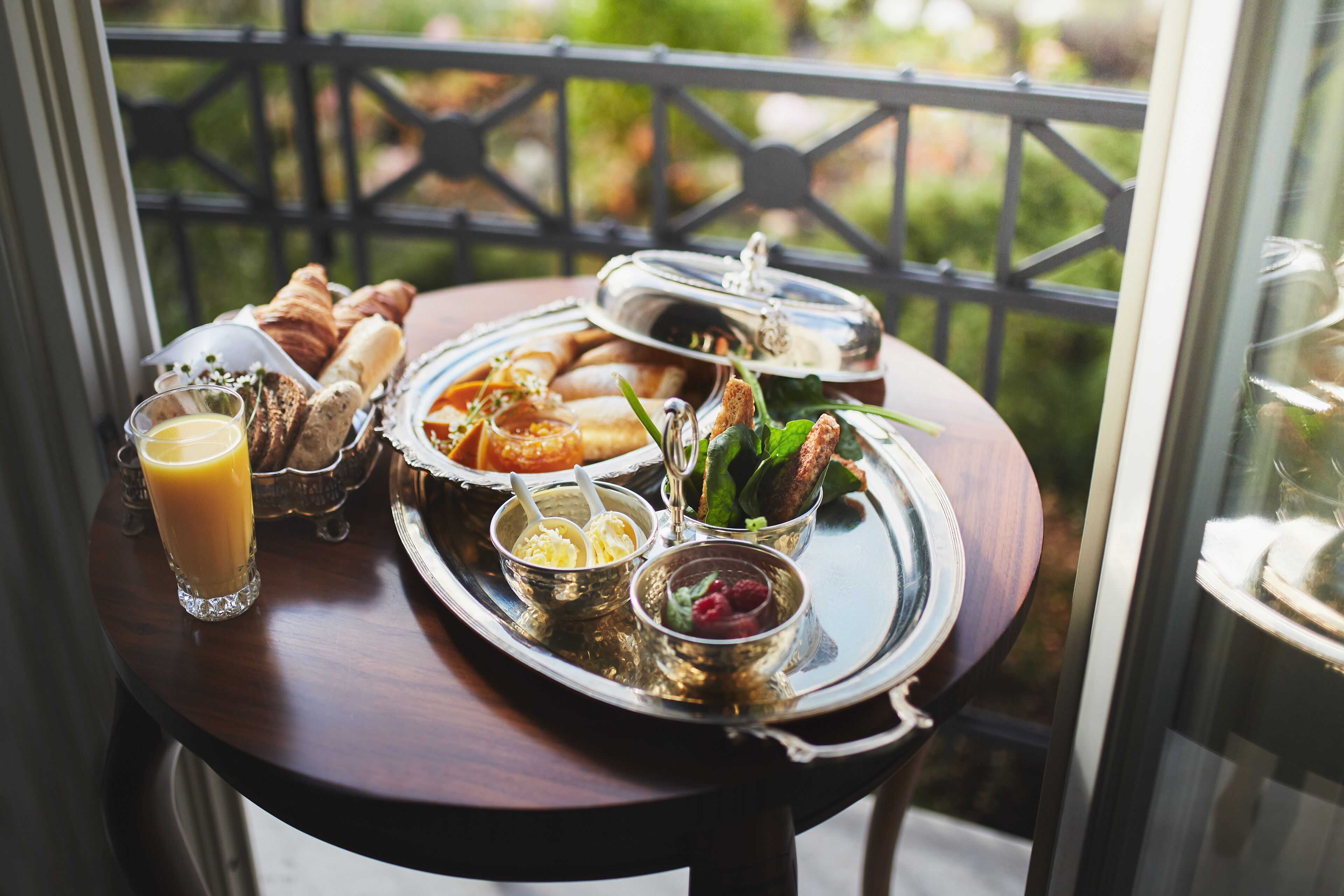 A breakfast tray with croissants, pastries, and a glass of orange juice on a balcony with a view of greenery.