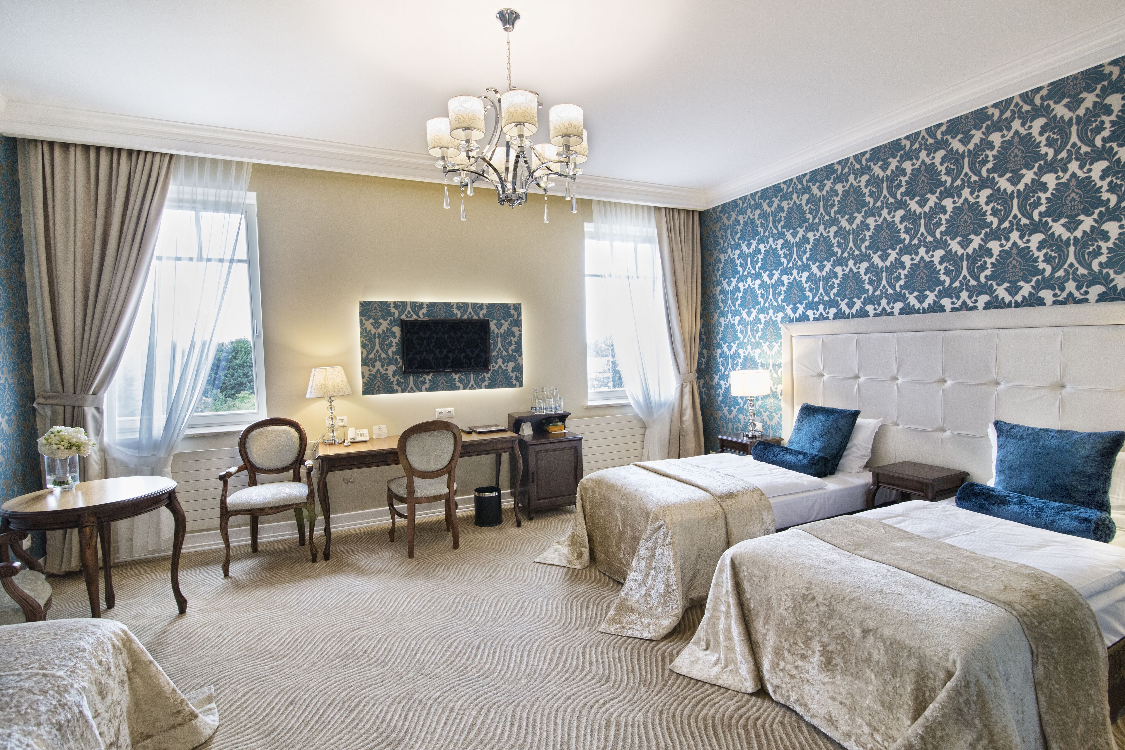 A hotel room with two beds, a desk, a chair, a TV, and a chandelier.