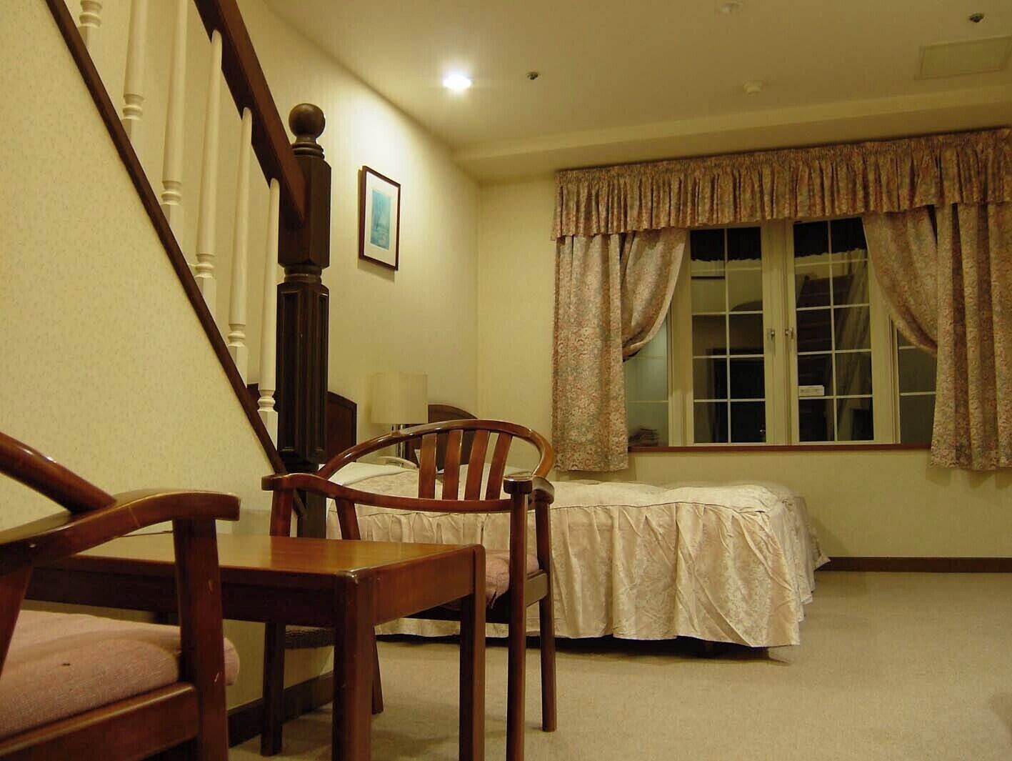 Un dormitorio con cama, una silla, una escalera de madera y una ventana con cortinas.