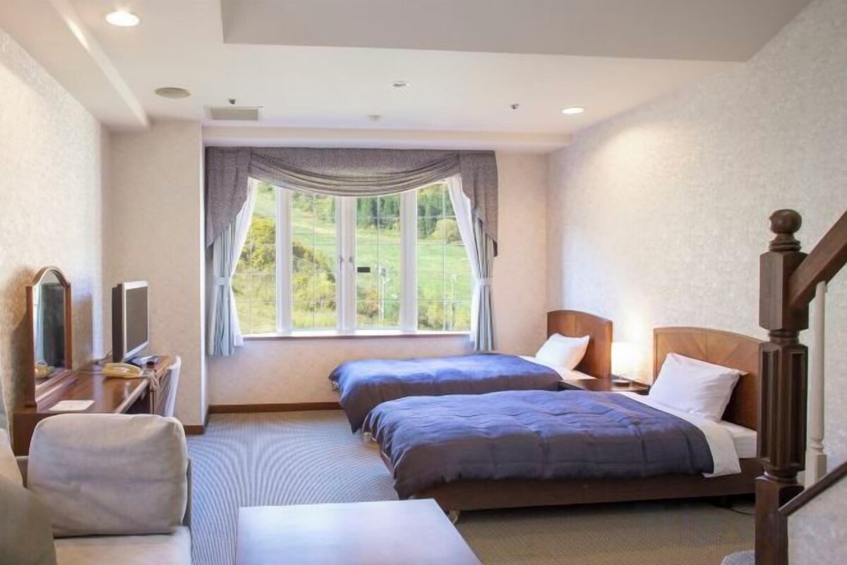 Ein Hotelzimmer mit zwei Betten, einem Schreibtisch mit Stuhl, einem Fernseher und einem Fenster mit Vorhängen.