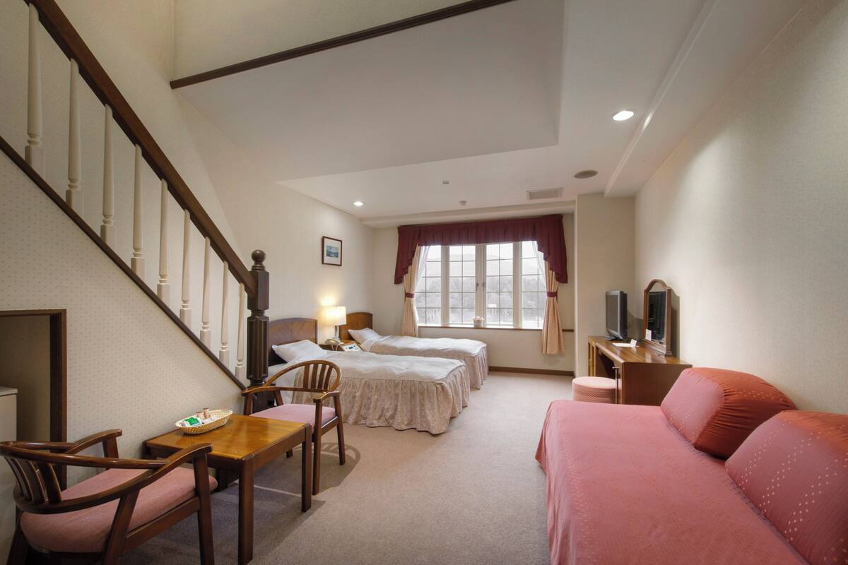 Habitación de hotel con escalera, dos camas, un escritorio con lámpara, televisor y sofá rojo.