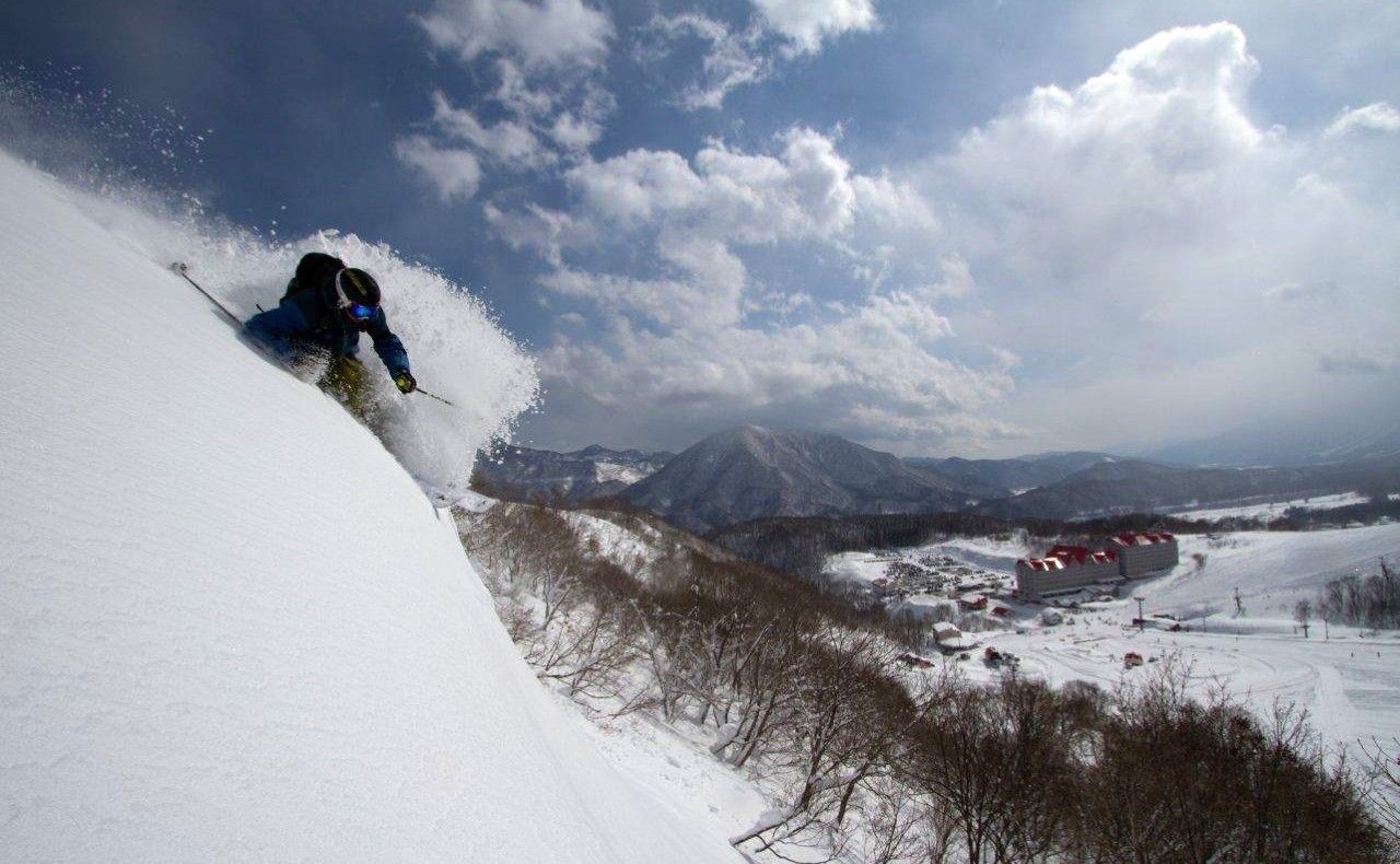 A skier on a snowy slope with a mountainous backdrop and a small settlement in the distance.