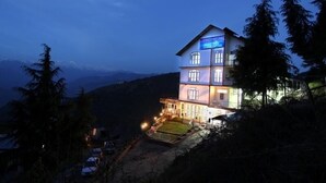 A multi-story hotel with a balcony and a lit sign, surrounded by trees and hills. - Shining Star Resort (Chamba)