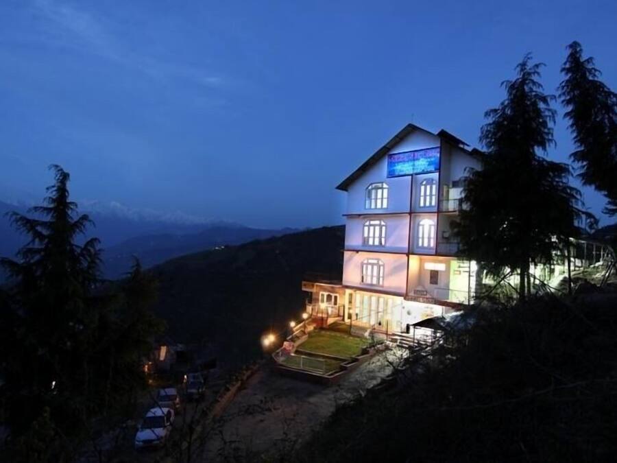 Un hotel de varios pisos con un balcón y un letrero iluminado, rodeado de árboles y colinas.
