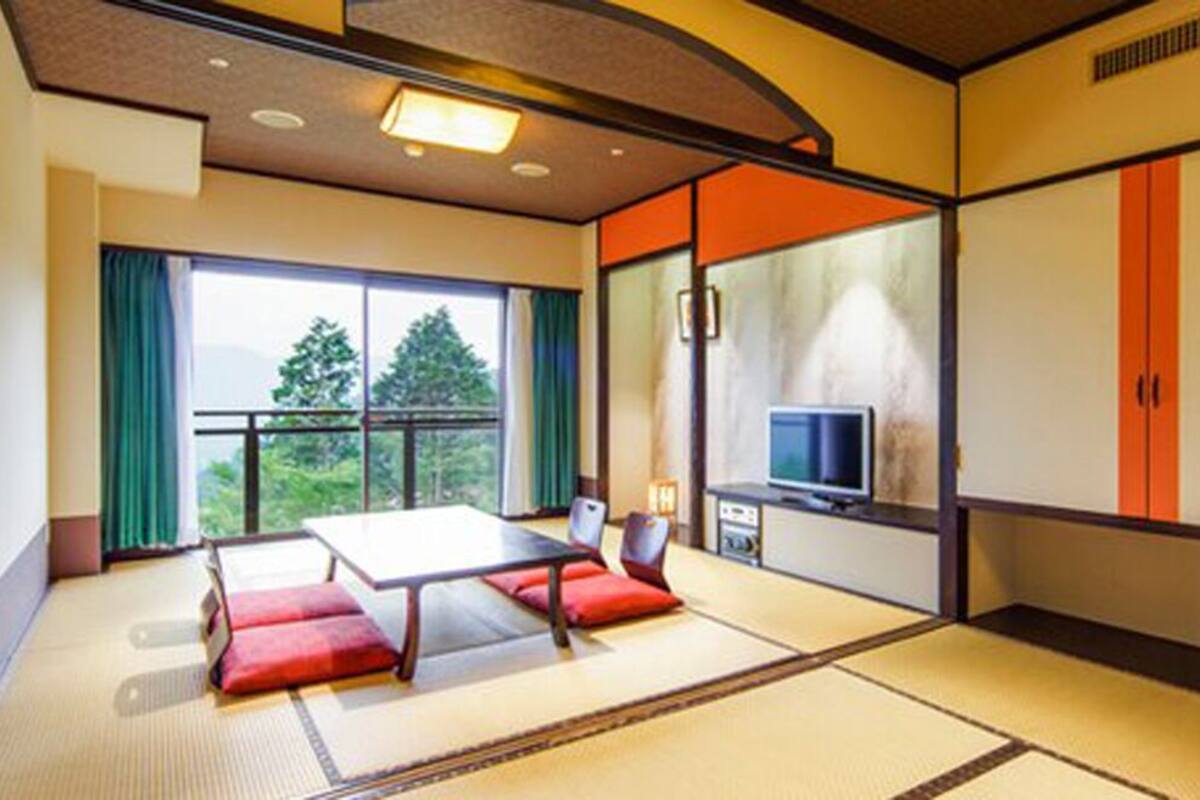 A room with tatami flooring, a low table with cushions, a television, and a sliding door leading to a balcony.