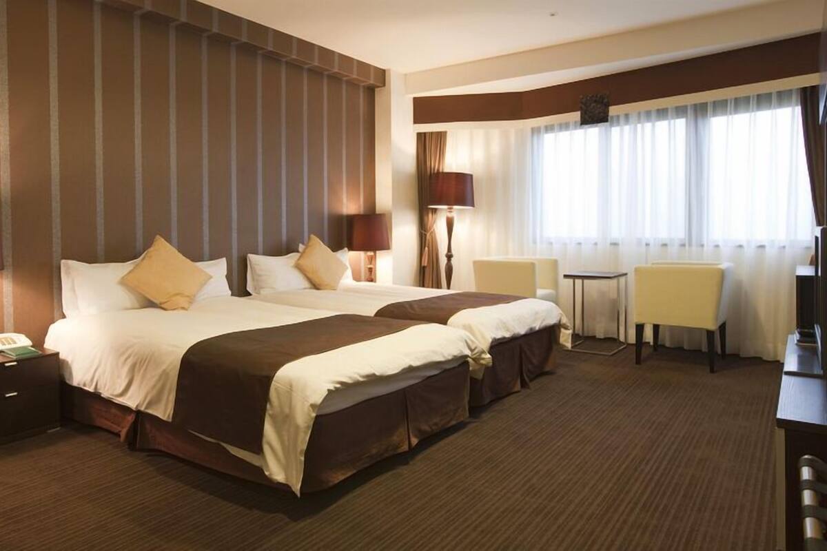 A hotel room with two beds, a desk, chairs, and a large window with curtains.