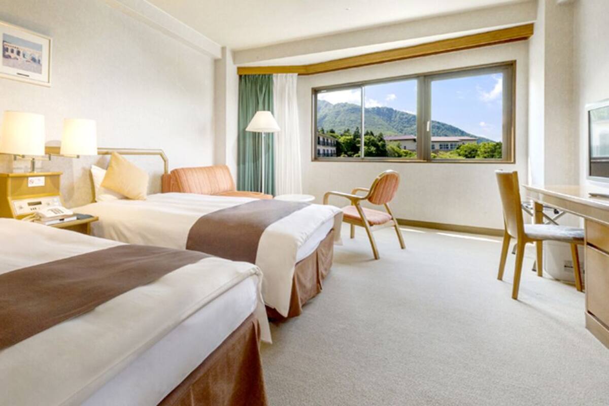 A hotel room with two beds, a desk, a chair, and a large window with a mountain view.