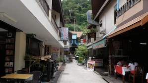 A narrow street with shops on either side, including a P&P House and a laundry service. There are outdoor seating areas and bicycles parked along the street.