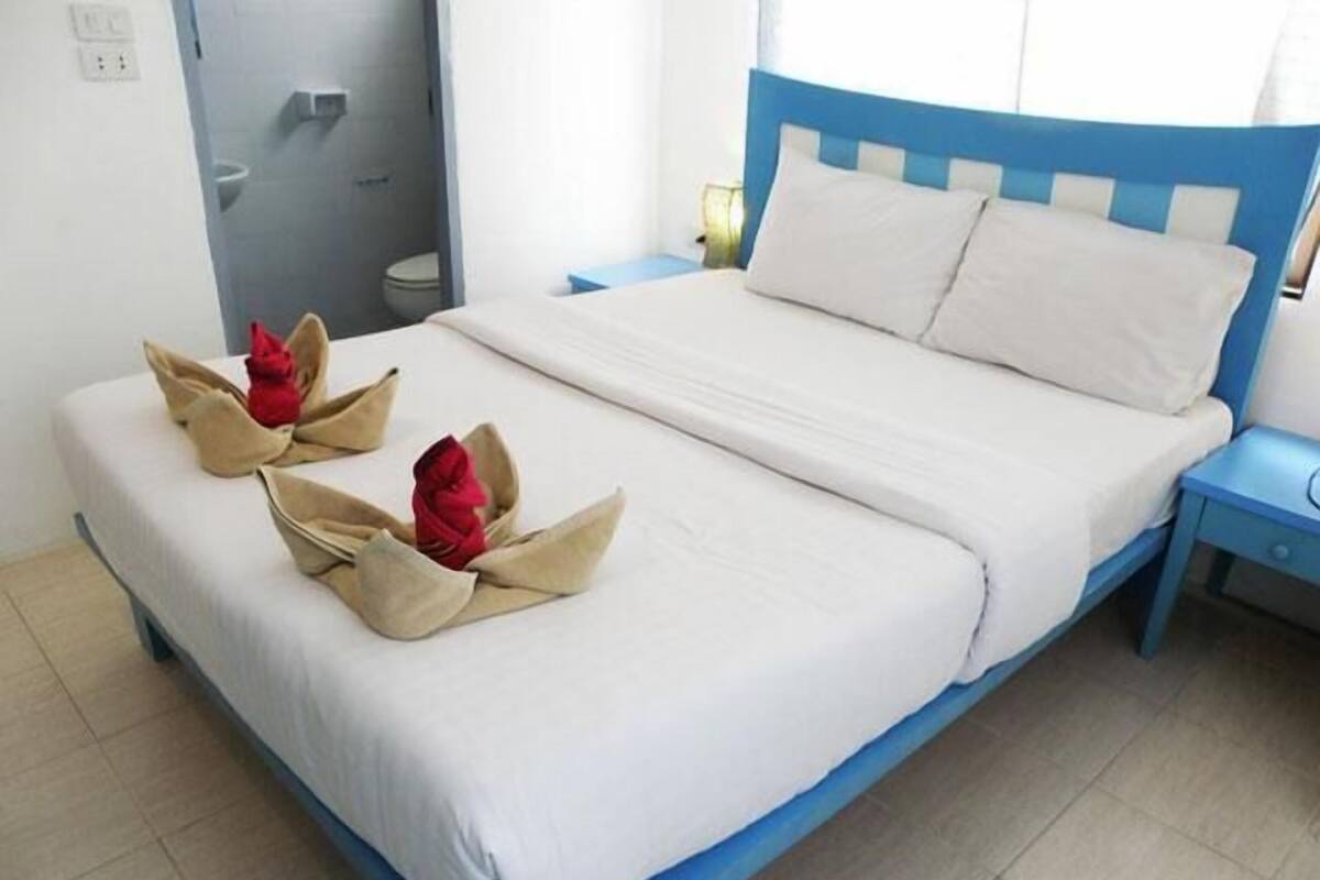 A neatly made bed with a blue headboard and white bedding, a small bedside table with a lamp, and a bathroom visible in the background.