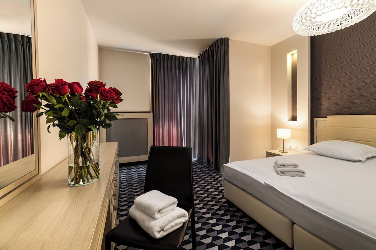 Quarto de hotel com cama, uma cadeira, escrivaninha com um vaso de flores, espelho, mesa de cabeceira com abajur e piso estampado.