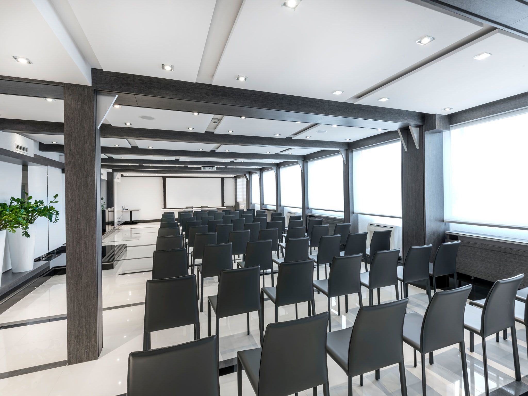 A modern conference room with rows of black chairs, a large screen, and a potted plant.