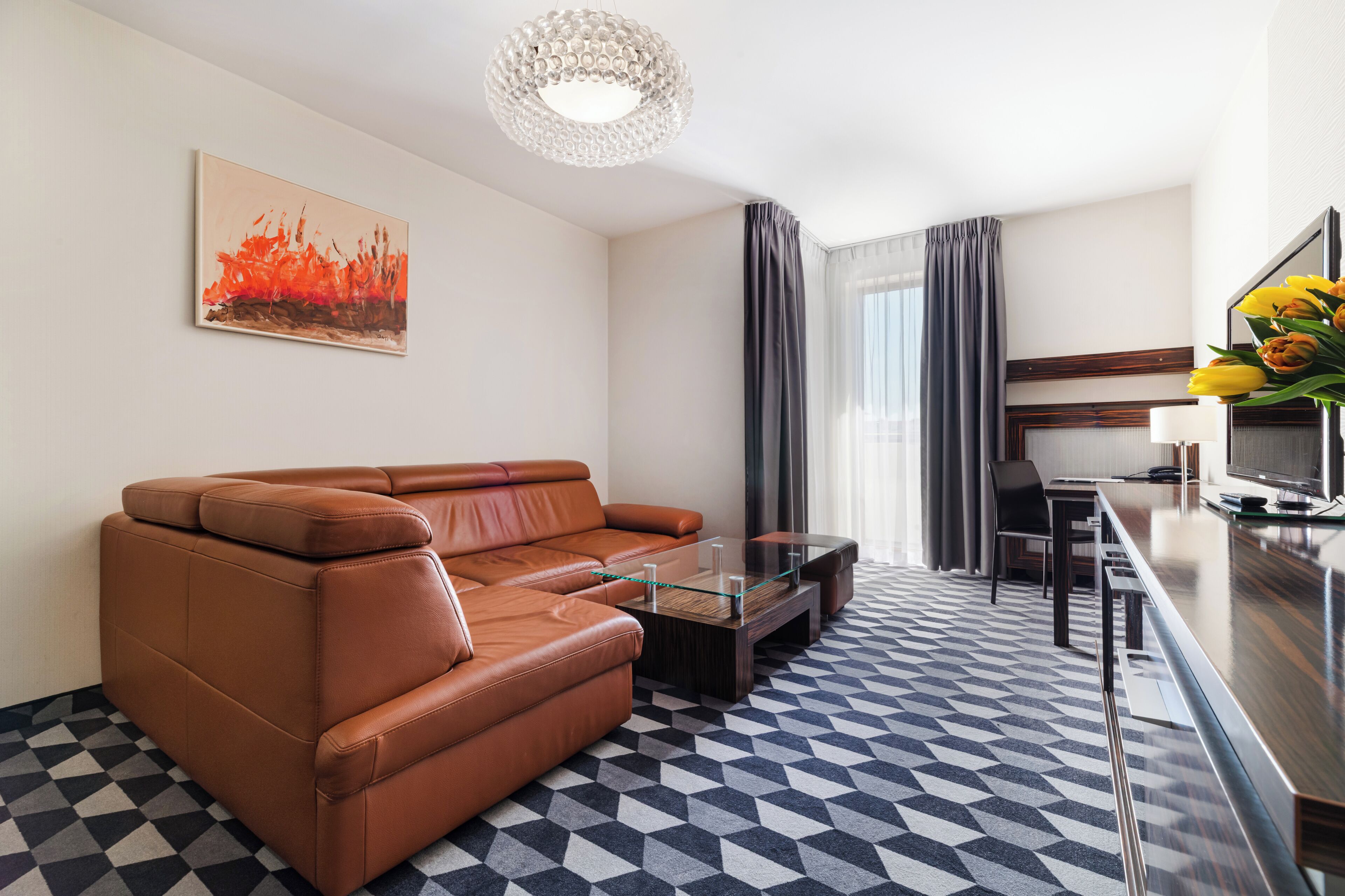 Una sala de estar moderna con un sofá de cuero marrón, una mesa de centro de vidrio, un comedor con una mesa larga y sillas, y un ventanal con cortinas.