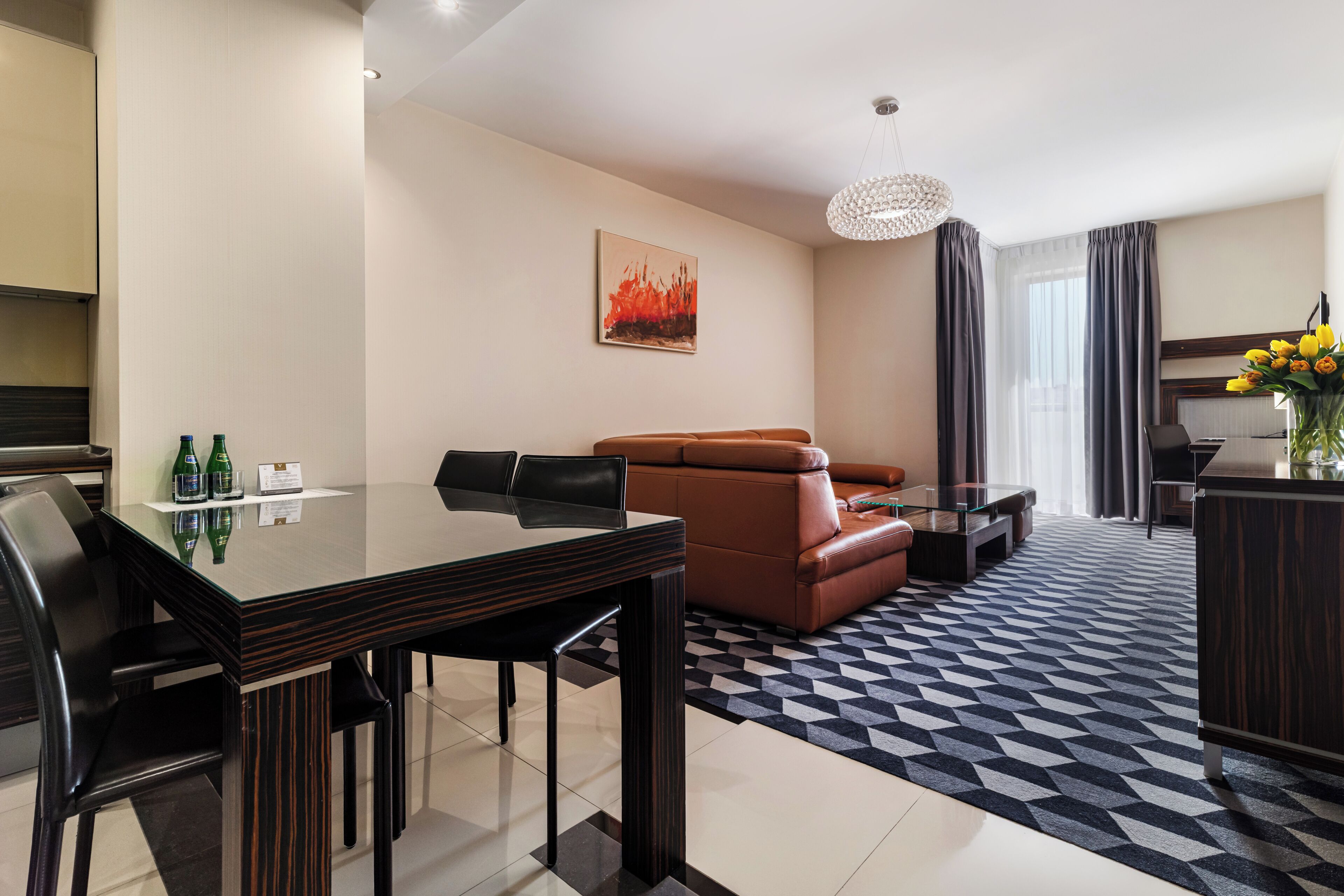 A modern hotel room with a dining table, leather sofa, and a chandelier.