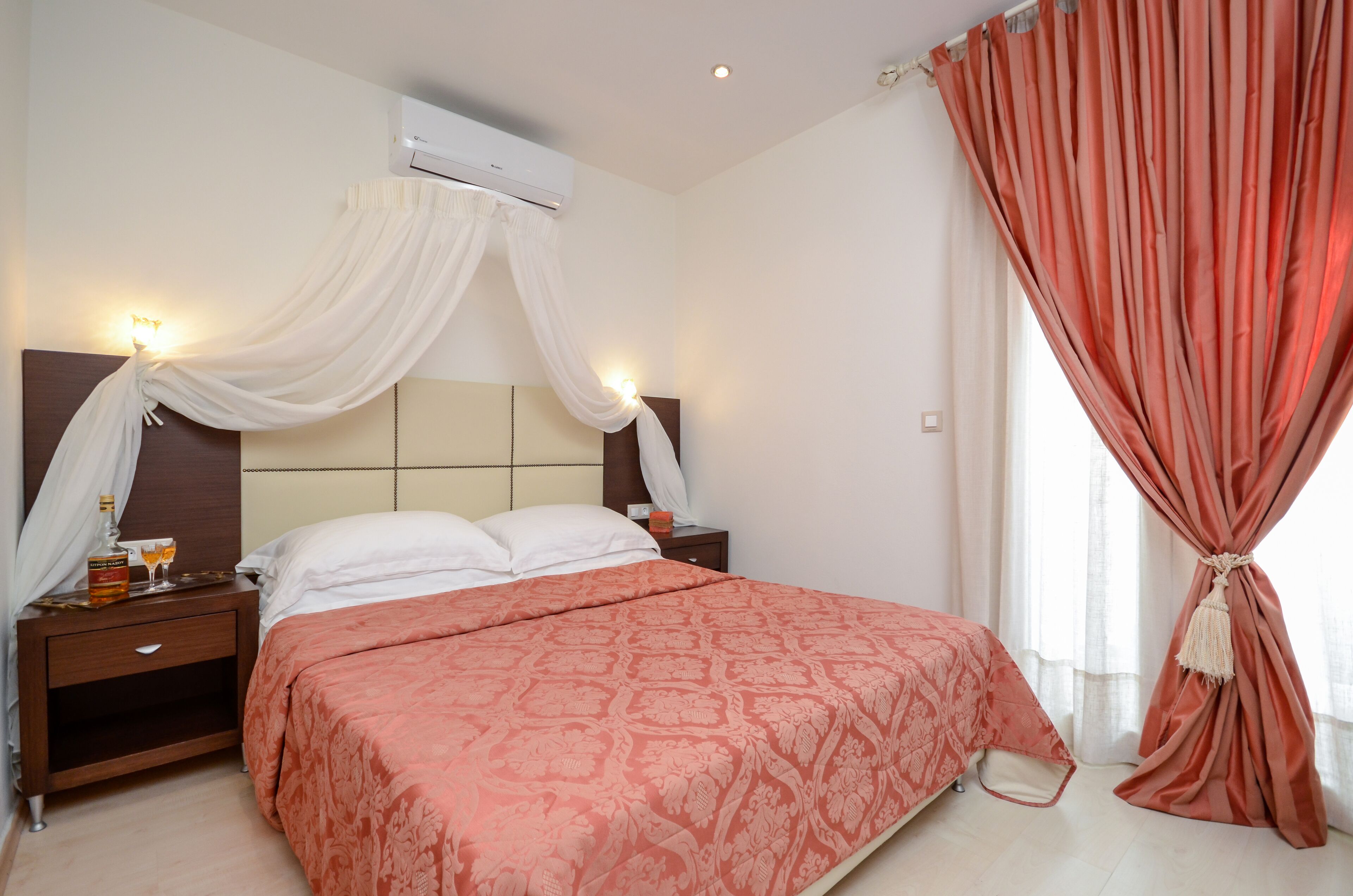 A neatly made bed with a patterned bedspread, a bedside table with a bottle and glasses, and a wall-mounted air conditioner.