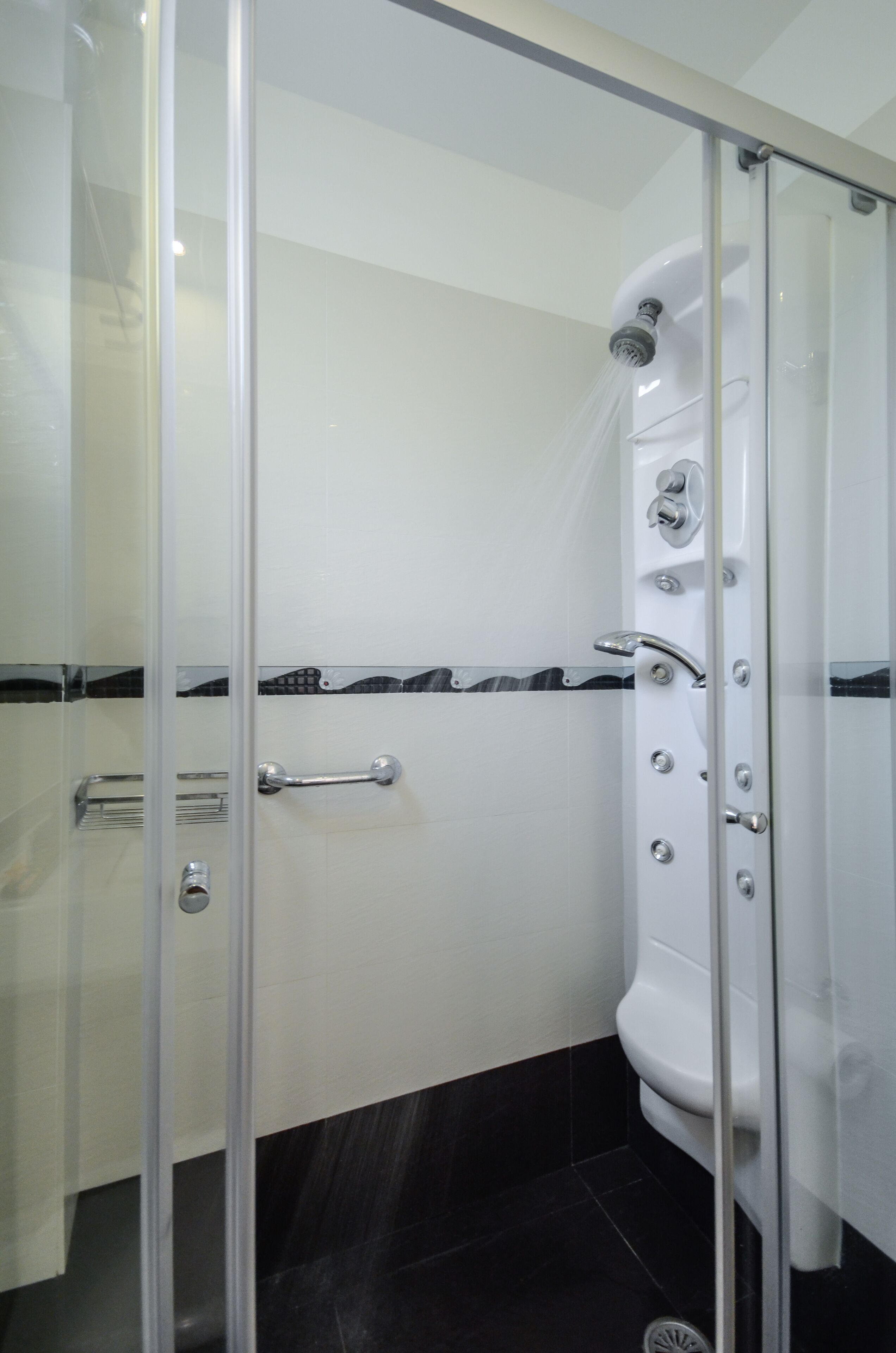 A glass-enclosed shower with a white tiled wall, a black and white tiled floor, and a chrome showerhead.