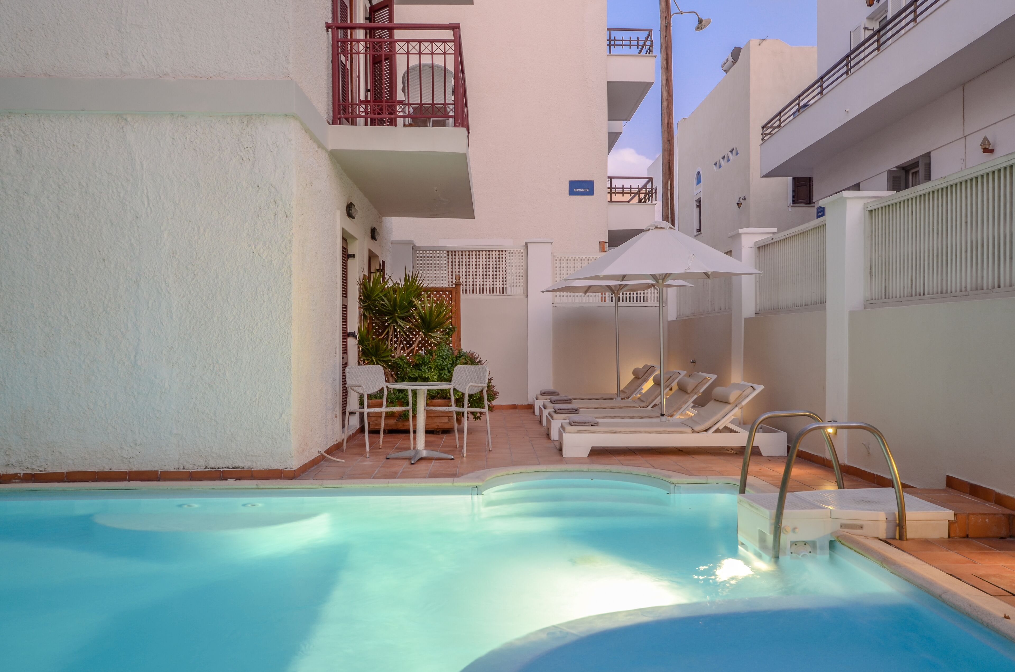 Una piscina con sillas de descanso y una mesa, rodeada por un edificio blanco con balcones.