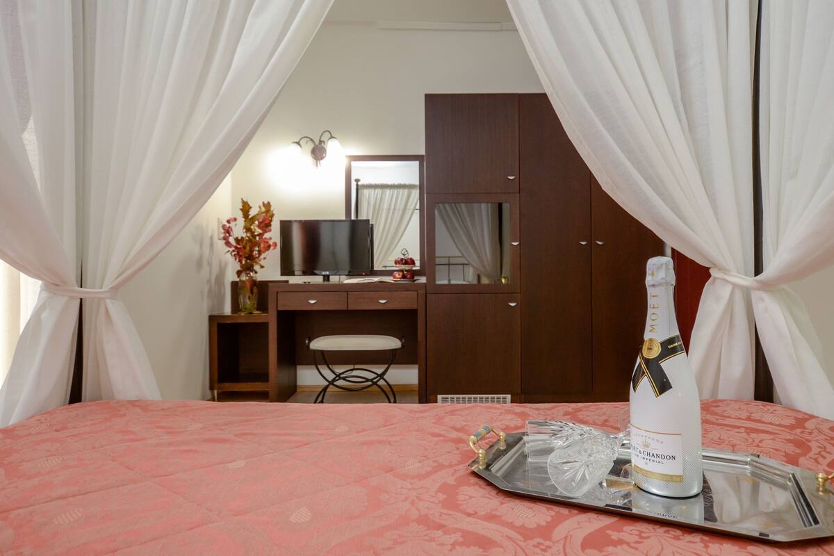 A hotel room with a bed, a desk, a chair, a television, a wardrobe, and a bottle of champagne on a tray.