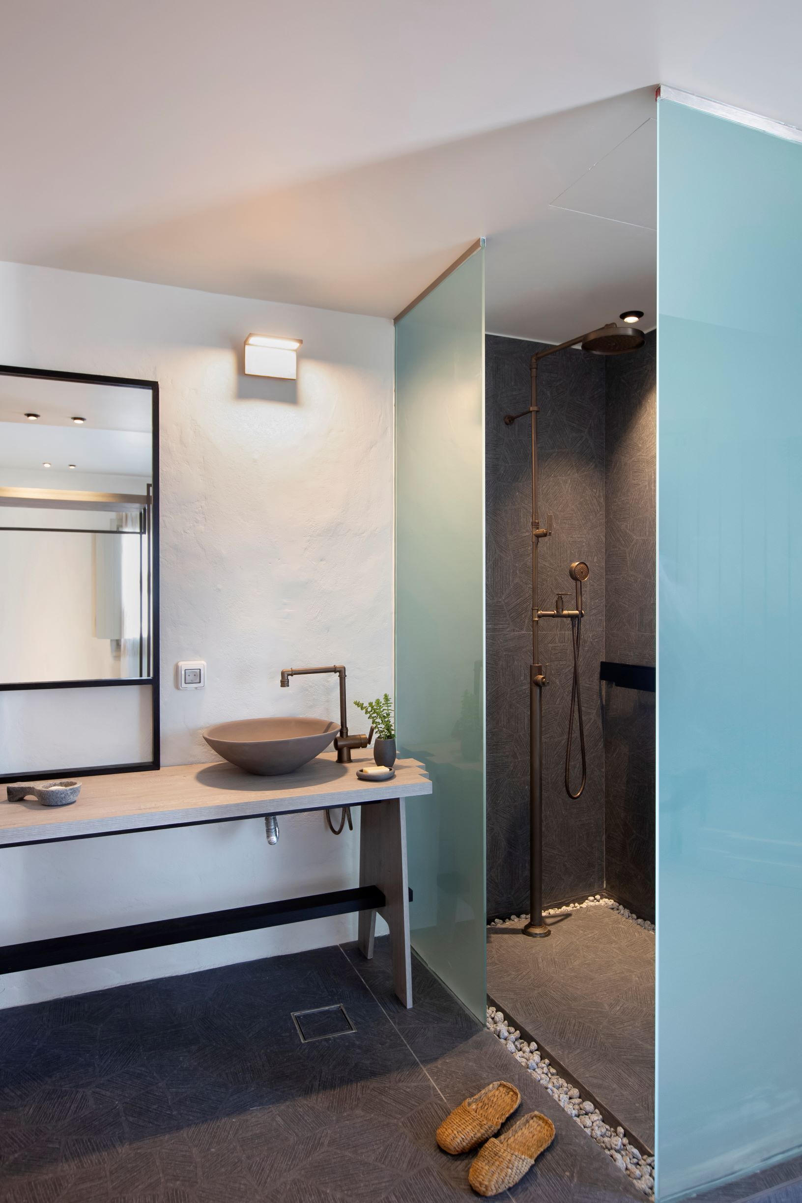 Een moderne badkamer met een grote spiegel, een wastafel op een voetstuk, een douche met een handdouche en een paar slippers op de vloer.