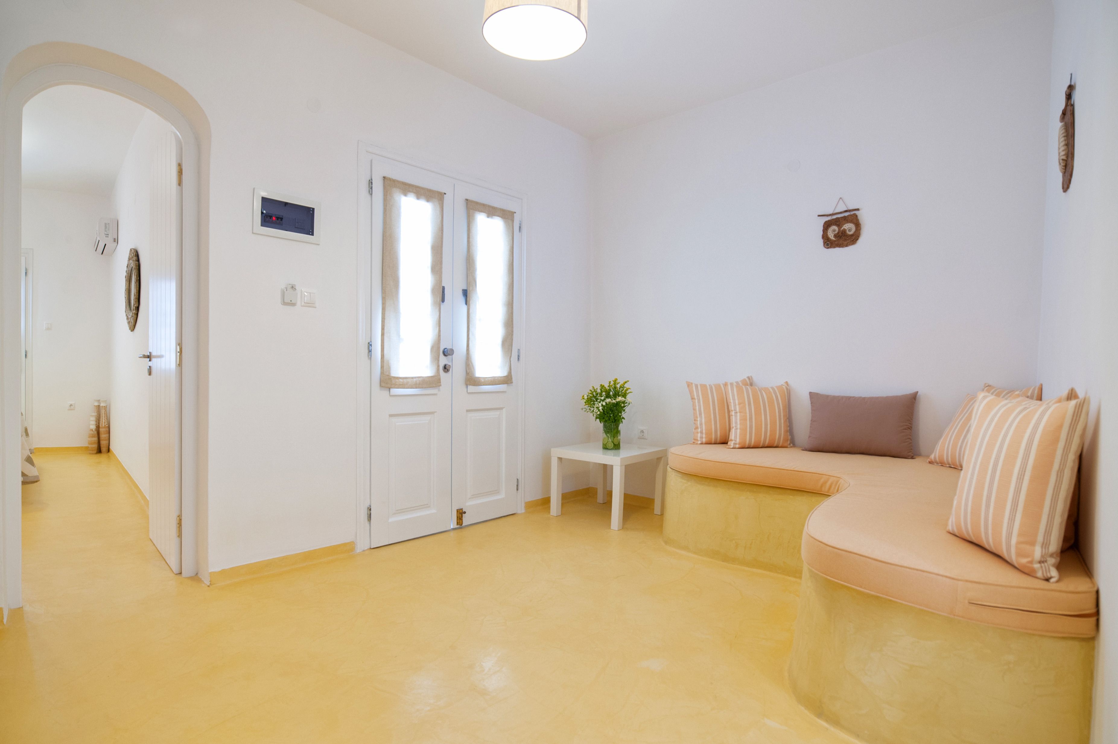 Una habitación luminosa con piso amarillo, una puerta blanca, una mesita con una planta y un sofá curvo con cojines.