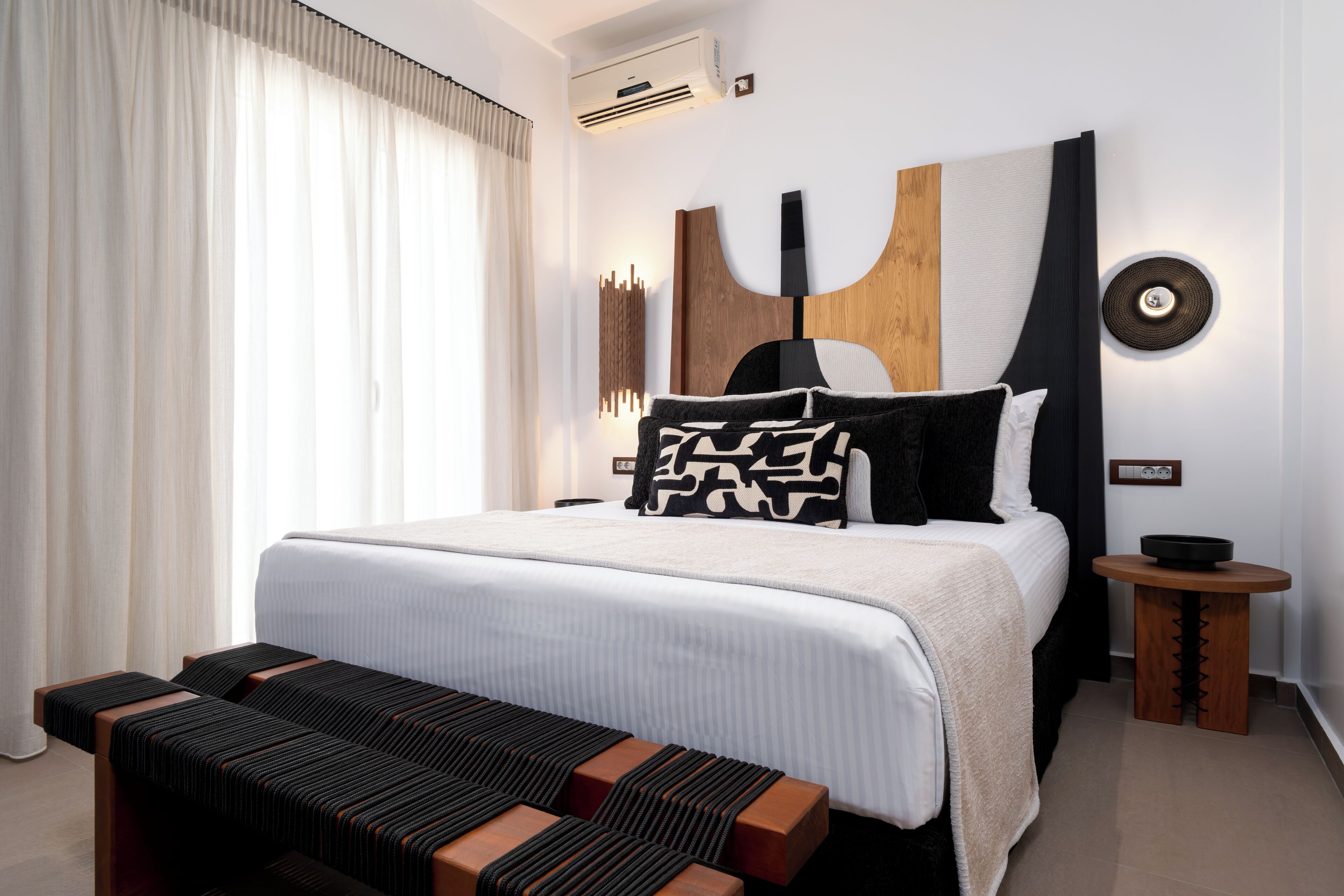 Een moderne hotelkamer met een groot bed, een nachtkastje, een bankje en een wandairconditioner.